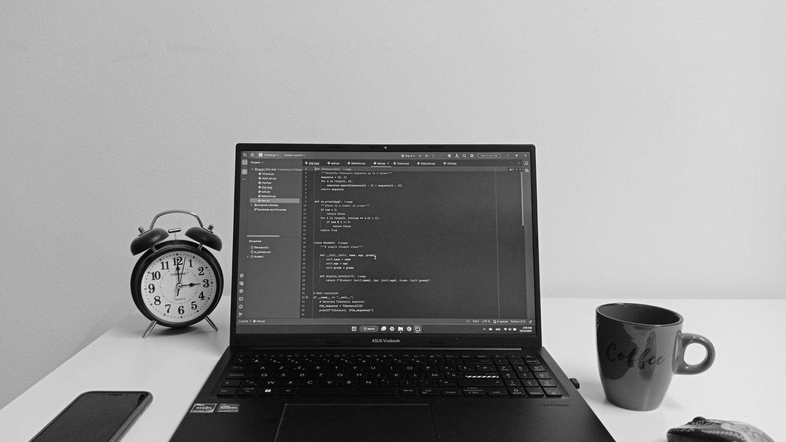 A monochrome desk with an open laptop displaying code, a phone, a clock, and a coffee mug